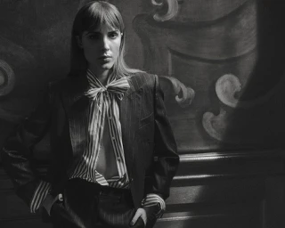 Freya Aspinall in a dark pinstripe suit and striped bow blouse posing against an ornate baroque wall mural.