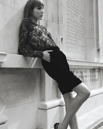 Model Freya Aspinall leaning against an inscribed stone wall wearing a sheer black lace long-sleeve top and tailored black shorts.