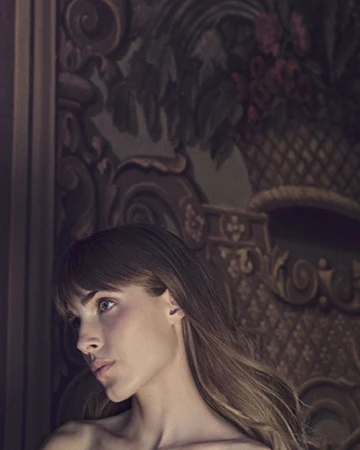 Profile portrait of a woman with bangs and bare shoulders in front of an ornate baroque floral wall backdrop.