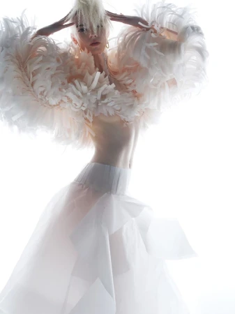 Anya Taylor-Joy wearing an exaggerated white feathered cape and tulle skirt against a bright white background.