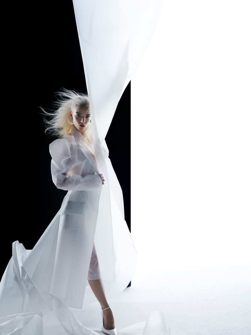 Anya Taylor-Joy posing in a sheer white sculpted coat amidst draped white fabric against a split black and white studio backdrop.