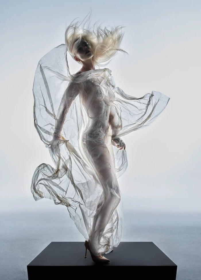 Anya Taylor-Joy posing on a black platform draped in sheer billowing plastic wrap against a light grey studio background.
