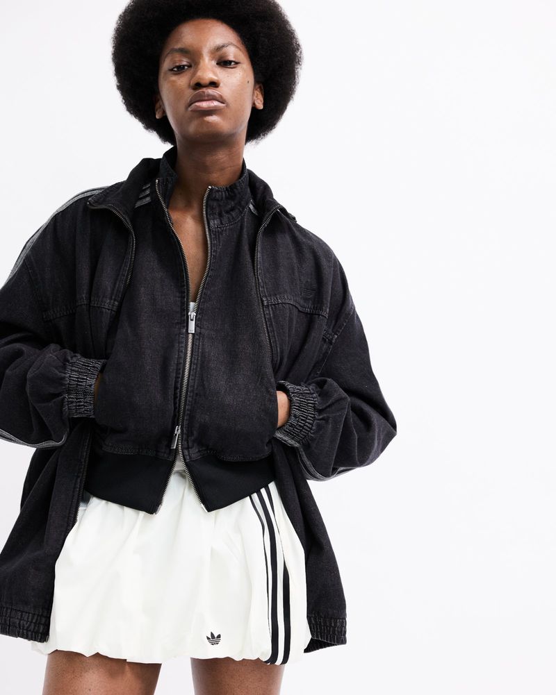 Black woman with an afro wearing a dark denim zip-up jacket and a white Adidas mini skirt against a white studio background.