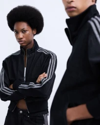 Grace Epolo wearing a black three-stripe zip track jacket with arms crossed alongside Luthando Ngema against a white backdrop.