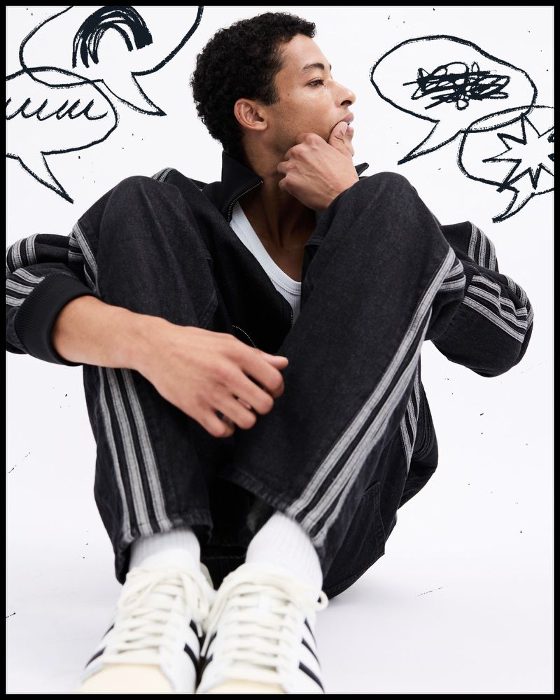 Man seated on a white studio floor wearing a dark striped denim tracksuit and white sneakers, with hand-drawn speech bubble doodles behind him.