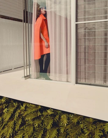Fashion model in a bright orange cape coat and black tights standing behind a sheer curtain inside a modern glass-walled house.