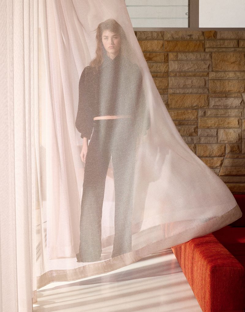 A model wearing black separates poses behind a translucent white curtain against a stone wall.
