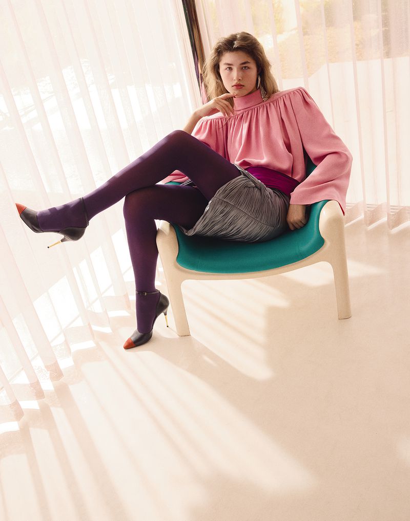 Model in a pink high-neck blouse and purple tights sitting with legs crossed on an armchair by sheer sunlit curtains.