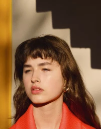 Close-up portrait of a woman with bangs in a coral collared top against a wall with a geometric stepped shadow.