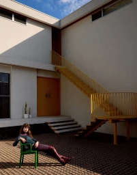 Model seated on a green chair in an architectural courtyard with a yellow exterior staircase and hard shadows.
