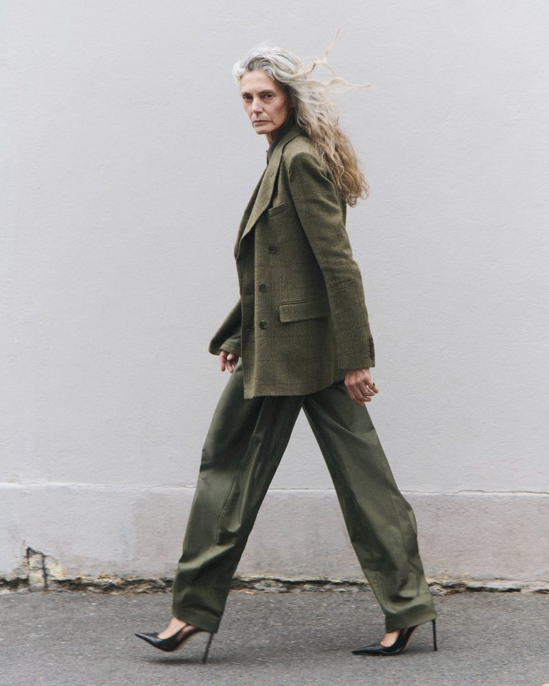 Model Sigrid de l'Epine walking in a green tailored suit and black stiletto heels against a plain urban exterior wall.