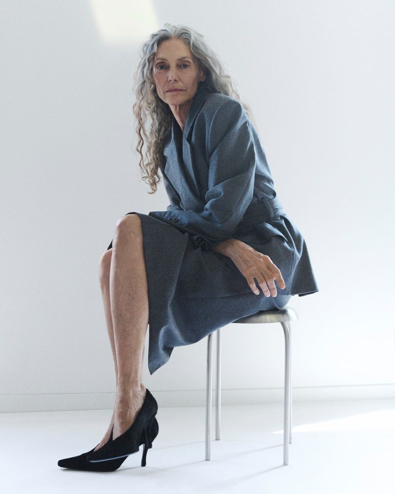 Model Sigrid de l'Epine seated on a metallic stool wearing a grey belted tailored coat and black pointed heels in a white studio.