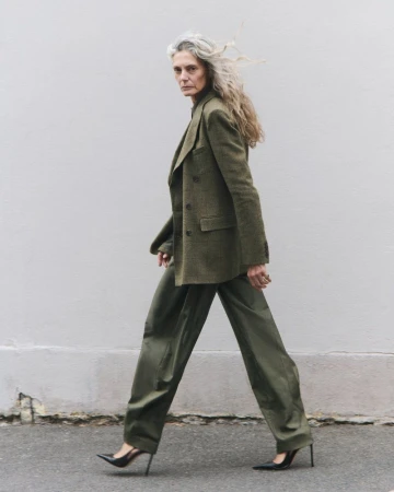 Model Sigrid de l'Epine walking in a green tailored suit and black stiletto heels against a plain urban exterior wall.