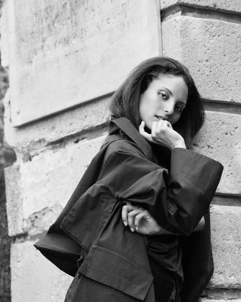 Monochrome portrait of model Michiko Foy wearing an oversized black coat, leaning against a stone building wall.