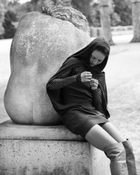 Model Michiko Foy wearing a dark hooded drape, mesh long sleeves, and tall boots while leaning against a large stone sculpture.