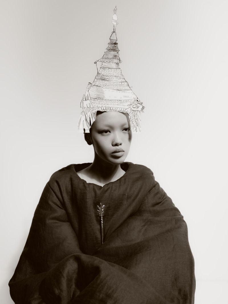 Model Weiyi Fang wearing an oversized dark draped cloak and an illustrated conical headpiece against a plain studio backdrop.