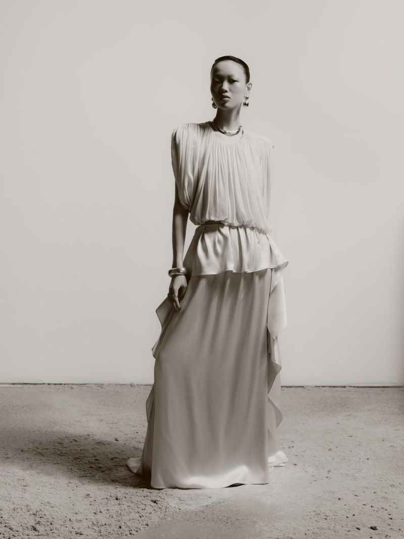 Model Weiyi Fang standing in a draped off-white sleeveless gown with silver jewelry against a neutral studio backdrop.