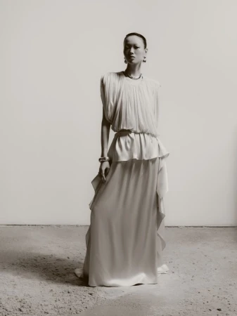 Model Weiyi Fang standing in a draped off-white sleeveless gown with silver jewelry against a neutral studio backdrop.