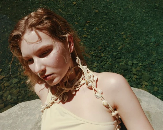 Model Anabel Krasnotsvetova wearing a seashell rope halter top while posing on a rock by river water in natural sunlight.