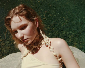 Model Anabel Krasnotsvetova wearing a seashell rope halter top while posing on a rock by river water in natural sunlight.