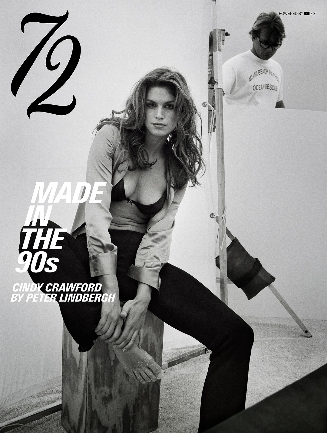 Cindy Crawford seated barefoot on a wooden apple box wearing an open shirt and black bra on an editorial photography set.