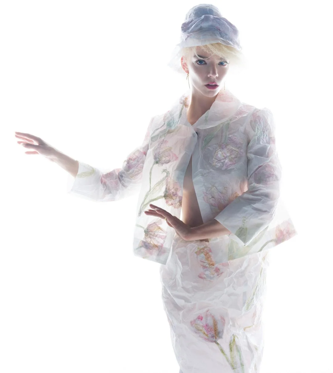 Anya Taylor-Joy posing in a sheer white floral printed jacket, matching skirt, and headpiece against a pure white background.