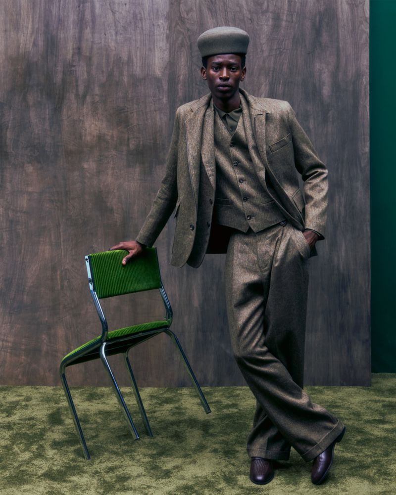 Model Adamu Bulus wearing a tailored three-piece suit and pillbox hat, resting his hand on a green chair.
