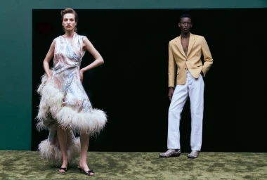 Diana Skowron in a feather-trimmed dress and Adamu Bulus in a yellow blazer posing on green carpet against a dark backdrop.