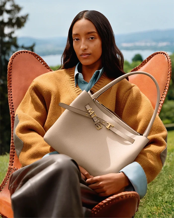 Model Mona Tougaard seated in a leather chair holding a beige Tod's handbag outdoors in front of a distant landscape view.