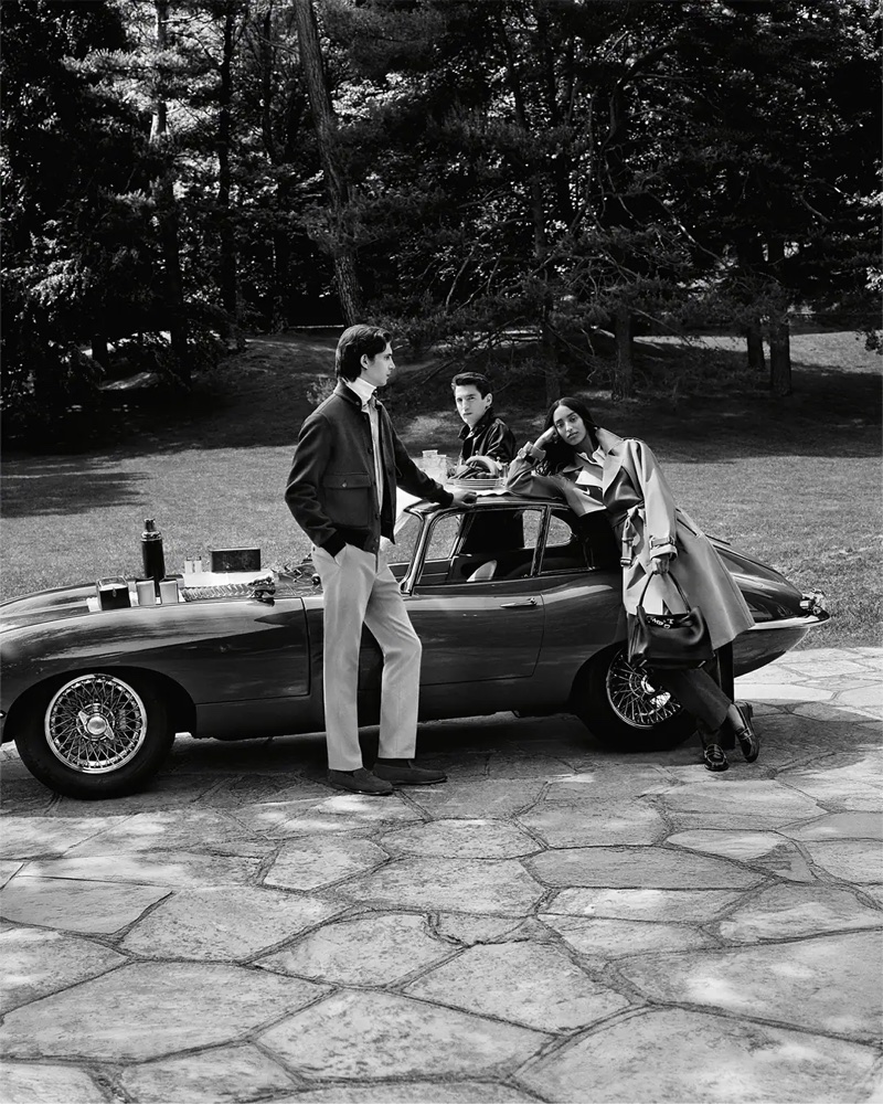 Monochrome fashion campaign image featuring models posing by a vintage sports car on a stone patio outdoors.