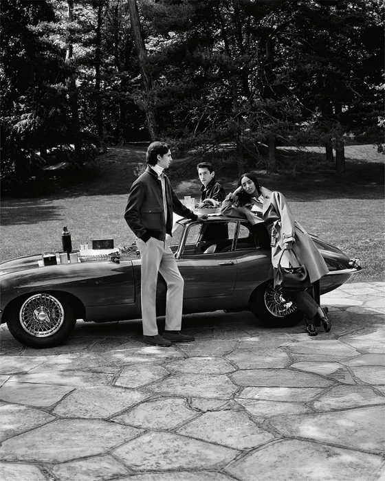 Monochrome fashion campaign image featuring models posing by a vintage sports car on a stone patio outdoors.