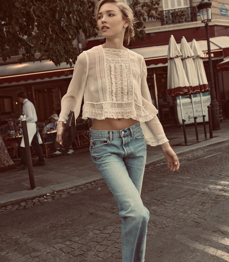 Model Thea Ros walking on a cobblestone street wearing a sheer cream lace cropped blouse and light wash denim jeans.