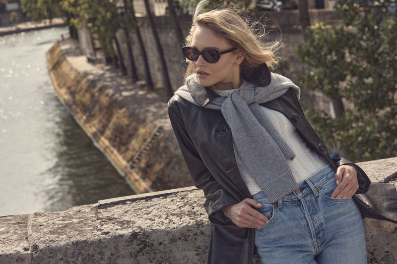Thea Ros wearing dark sunglasses, a black leather coat, a grey sweater, and blue denim jeans leaning against a stone bridge wall.