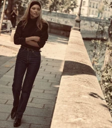 Model Thea Ros leaning against a stone embankment wall wearing dark denim straight-leg jeans, a black turtleneck, and a headband.