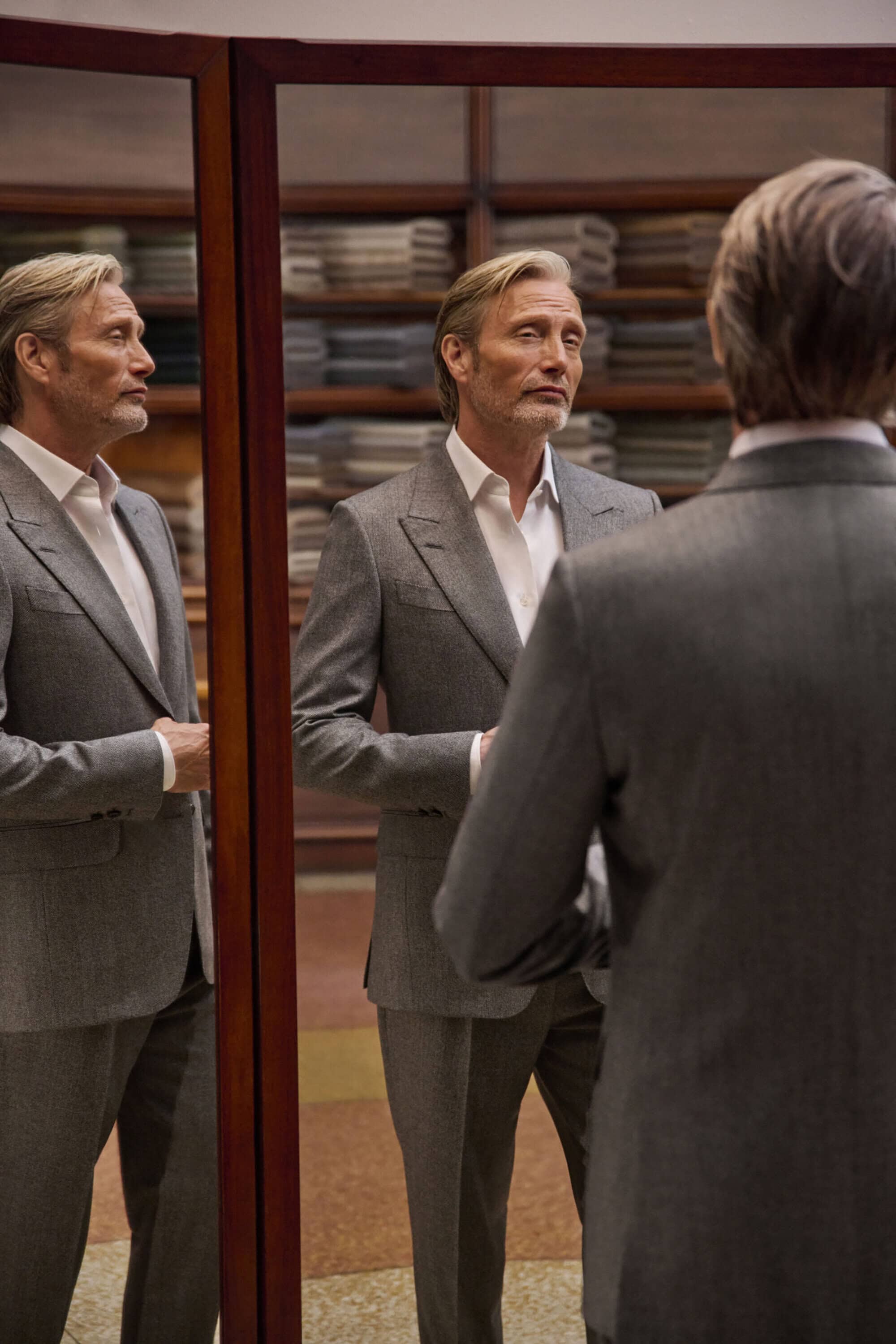 Mads Mikkelsen wearing a tailored grey suit and white shirt, looking at his reflection in a three-panel mirror in a tailor shop.