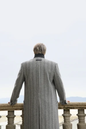 Mads Mikkelsen seen from behind wearing a grey pinstripe overcoat and leaning against a stone balustrade outdoors.