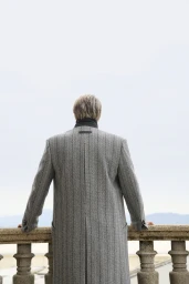 Mads Mikkelsen viewed from behind in a grey pinstripe overcoat leaning on a stone balustrade overlooking a misty horizon.