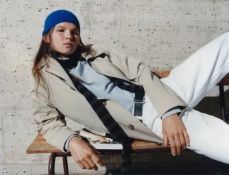 Model Georgia Martin wearing a beige trench coat, blue beanie, and white trousers reclining on a wooden bench against a stone wall.