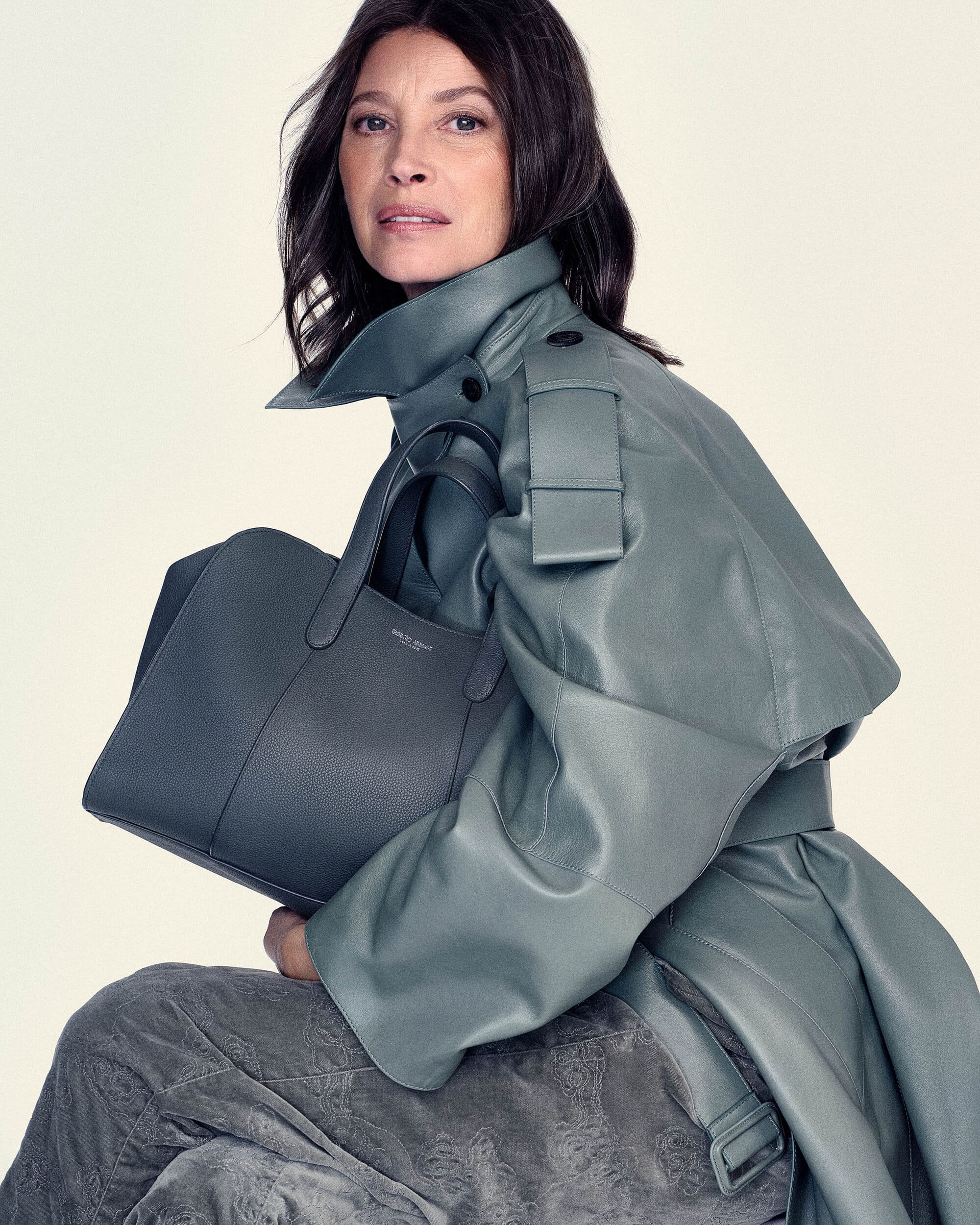 Christy Turlington wearing an oversized sage green leather trench coat and holding a structured handbag against a white studio backdrop.