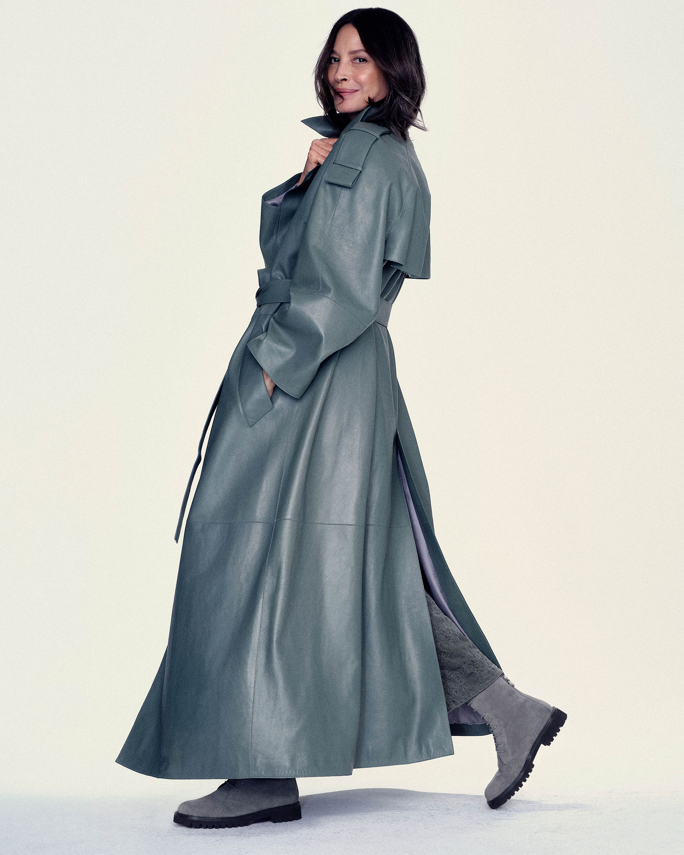 Christy Turlington poses looking back in a floor-length teal leather trench coat and grey boots against a white studio backdrop.