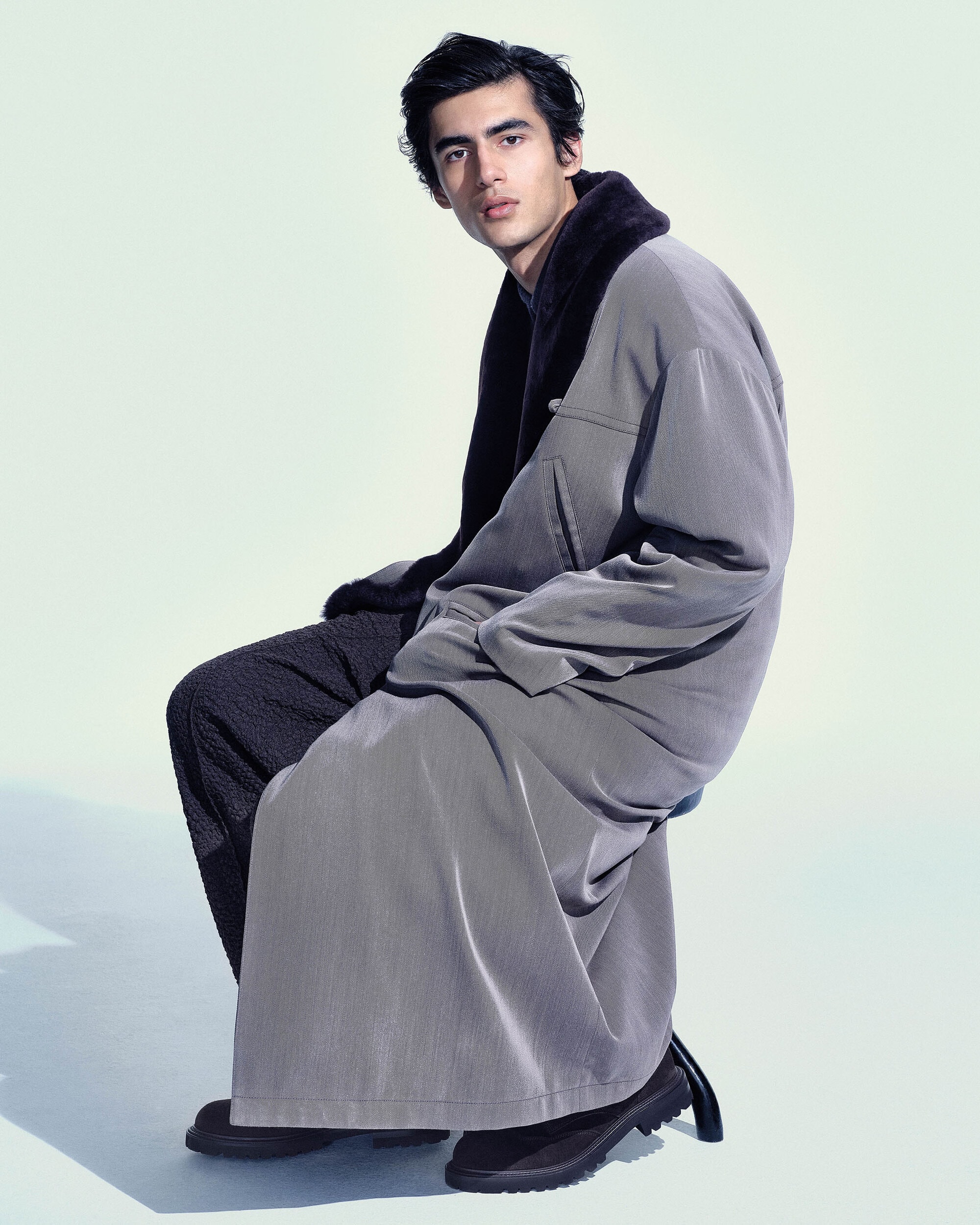 Model Akbar Shamji seated wearing an oversized grey overcoat with a dark collar in a studio setting for Giorgio Armani.