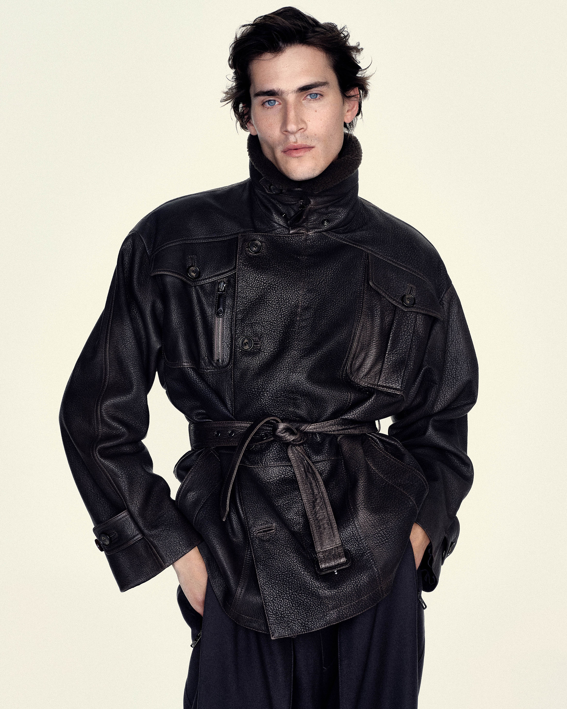 Male model in a textured black belted leather field jacket and dark trousers standing against a pale cream studio background.