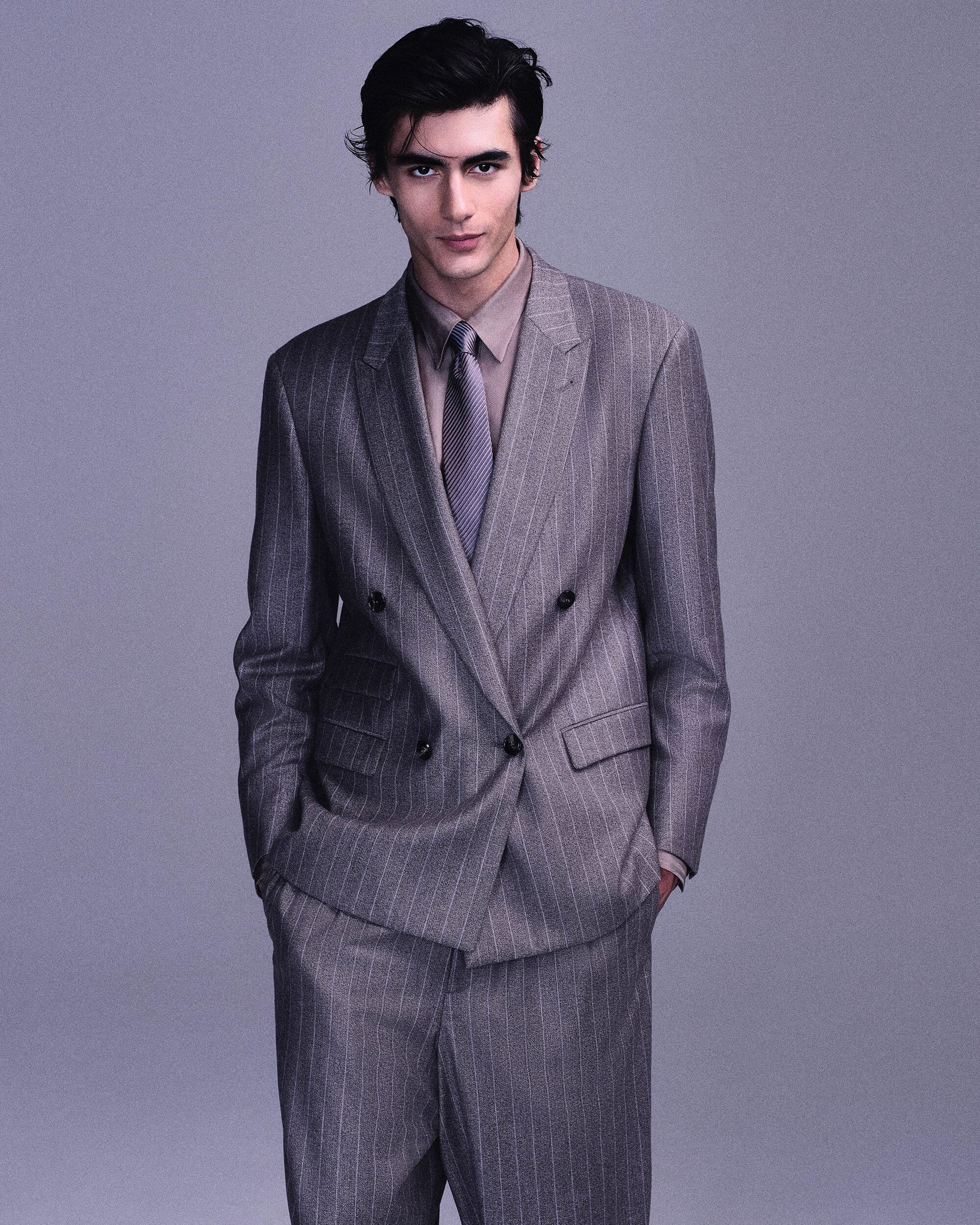 Model posing in a grey pinstriped double-breasted suit with a collared shirt and tie against a plain studio backdrop.