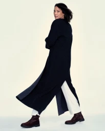 Christy Turlington wearing a long navy maxi coat, white trousers, and lace-up boots against a light studio backdrop.