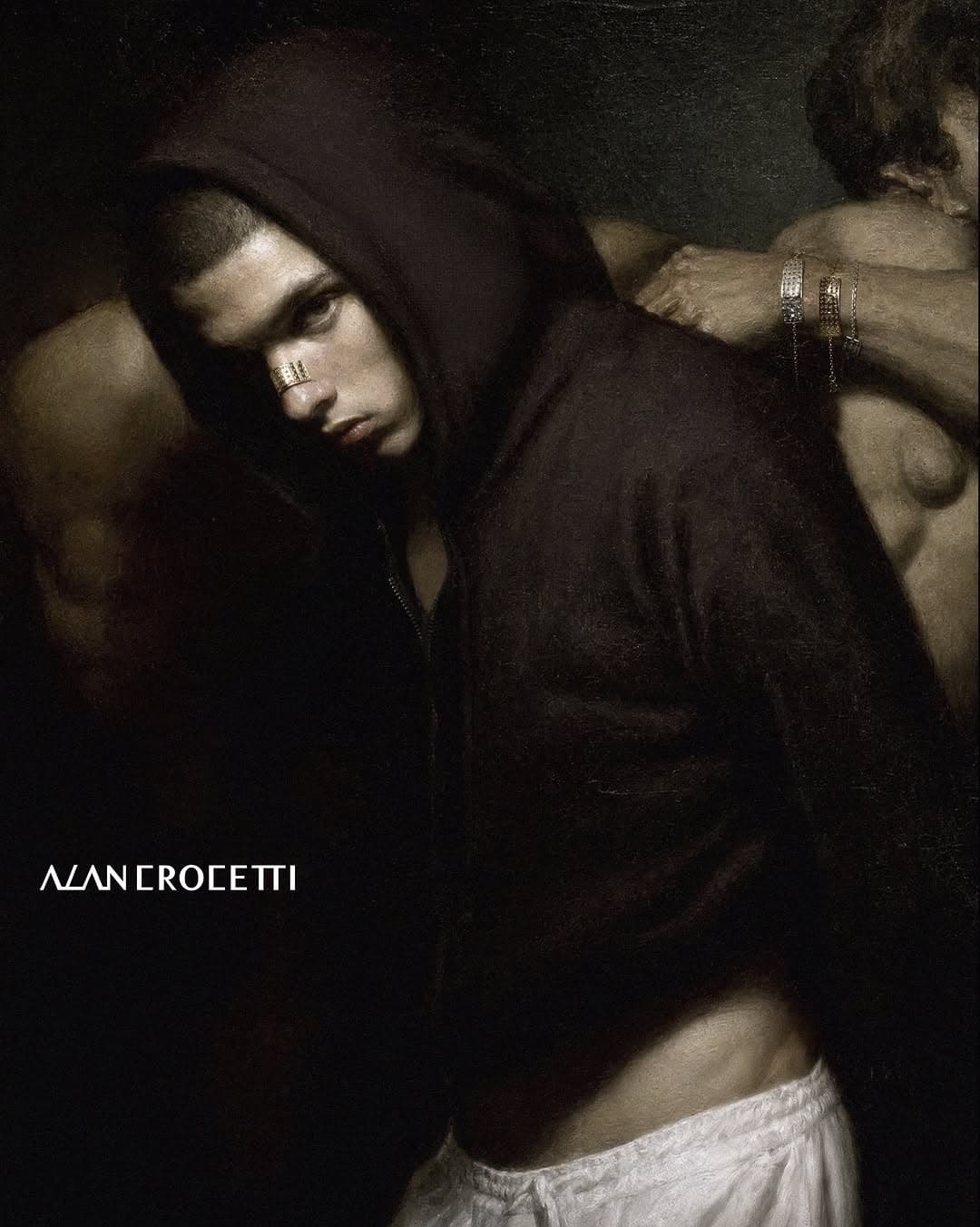 A man wearing a black hooded jacket and a silver metallic nose cuff against a dark chiaroscuro background.