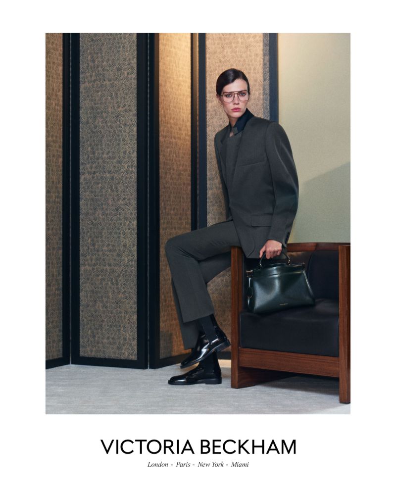 Model Alix Bouthors wearing a dark tailored suit and glasses, seated on an armchair holding a structured leather handbag.