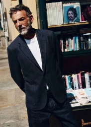 Alain Gossuin wearing a dark navy blazer, white t-shirt, and denim jeans standing in front of an outdoor bookstand in direct sunlight.