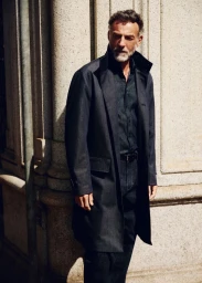 Model Alain Gossuin wearing a tailored dark denim overcoat and matching shirt standing against a stone architectural pillar.