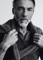 Alain Gossuin wearing a knit polo and sweater draped over his shoulders in a black and white portrait against a plain backdrop.