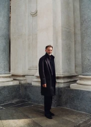 Alain Gossuin stands in a full-length black overcoat beside classical stone columns in soft natural daylight.